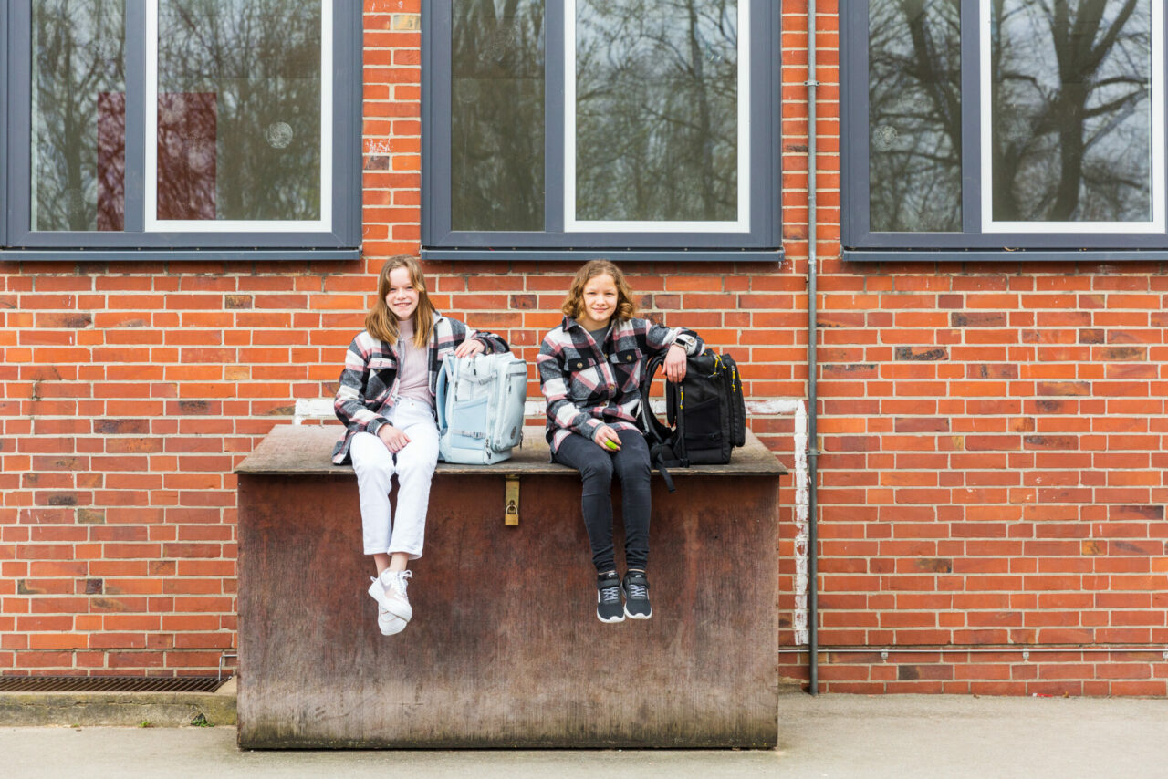 Die Zwillinge Lisa und Nele sitzen mit ihrem Schulrucksack von Kattbjoern vor einer Ziegelwand